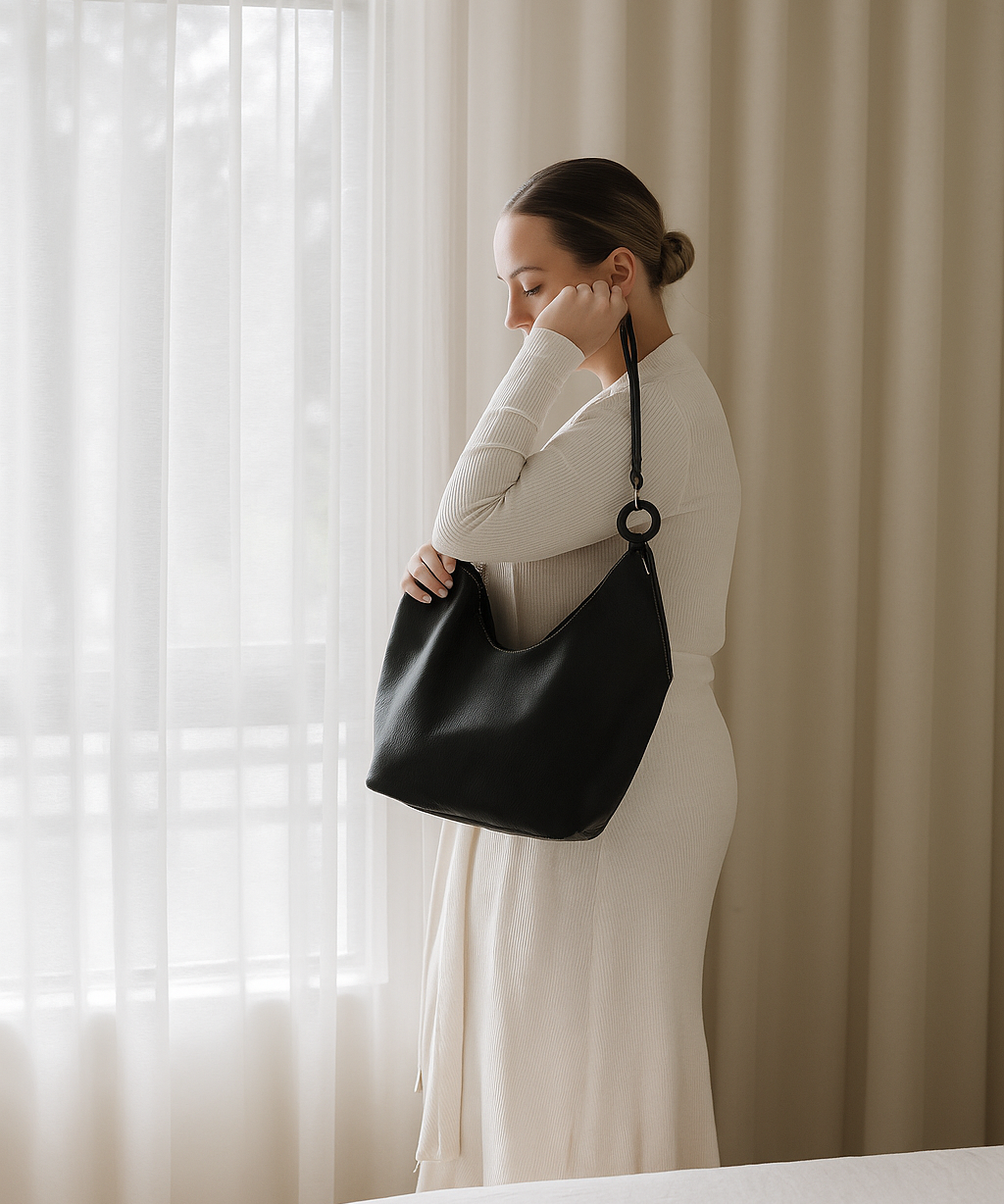 Woman holding a black ovae handbag in front of a window with white curtains