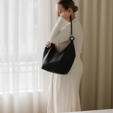 Woman holding a black ovae handbag in front of a window with white curtains