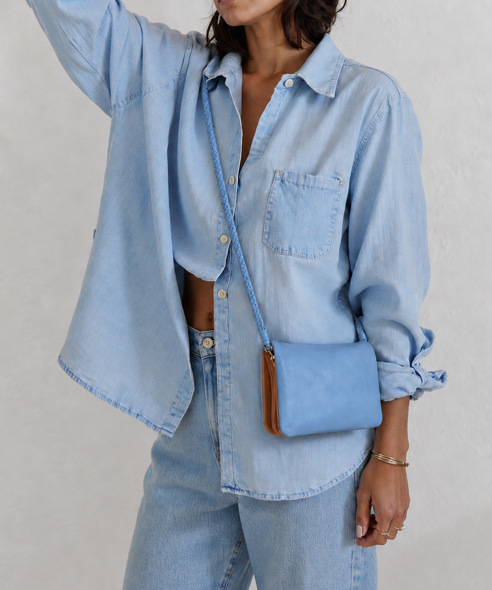 A woman wearing a light blue denim shirt and matching jeans, carrying a small light blue crossbody bag with a brown leather trim against a textured off-white wall.
