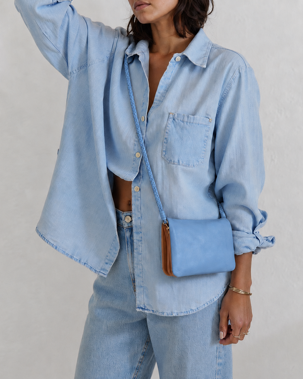 A woman wearing a light blue denim shirt and matching jeans, carrying a small light blue crossbody bag with a brown leather trim against a textured off-white wall.