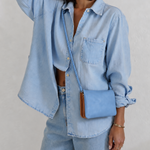 A woman wearing a light blue denim shirt and matching jeans, carrying a small light blue crossbody bag with a brown leather trim against a textured off-white wall.
