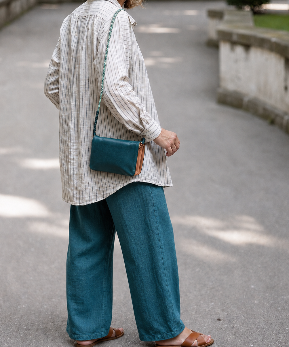 A person standing outdoors on a paved path, shown from the back and wearing a light striped shirt, teal wide-leg pants, and brown sandals, with a teal crossbody bag slung over the shoulder.