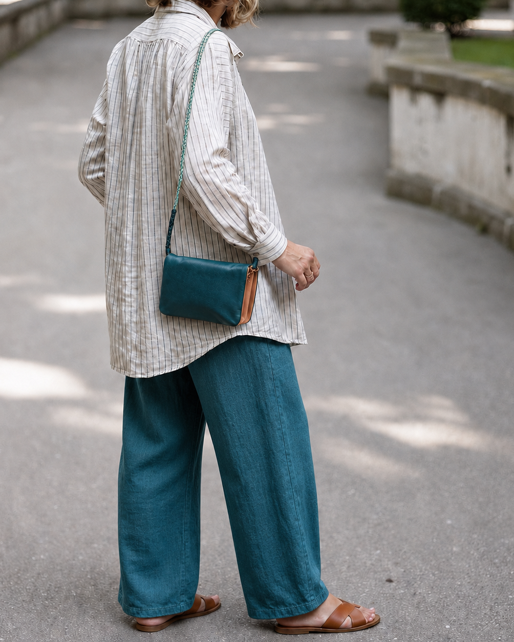 A person standing outdoors on a paved path, shown from the back and wearing a light striped shirt, teal wide-leg pants, and brown sandals, with a teal crossbody bag slung over the shoulder.