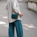 A person standing outdoors on a paved path, shown from the back and wearing a light striped shirt, teal wide-leg pants, and brown sandals, with a teal crossbody bag slung over the shoulder.