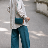 A person standing outdoors on a paved path, shown from the back and wearing a light striped shirt, teal wide-leg pants, and brown sandals, with a teal crossbody bag slung over the shoulder.