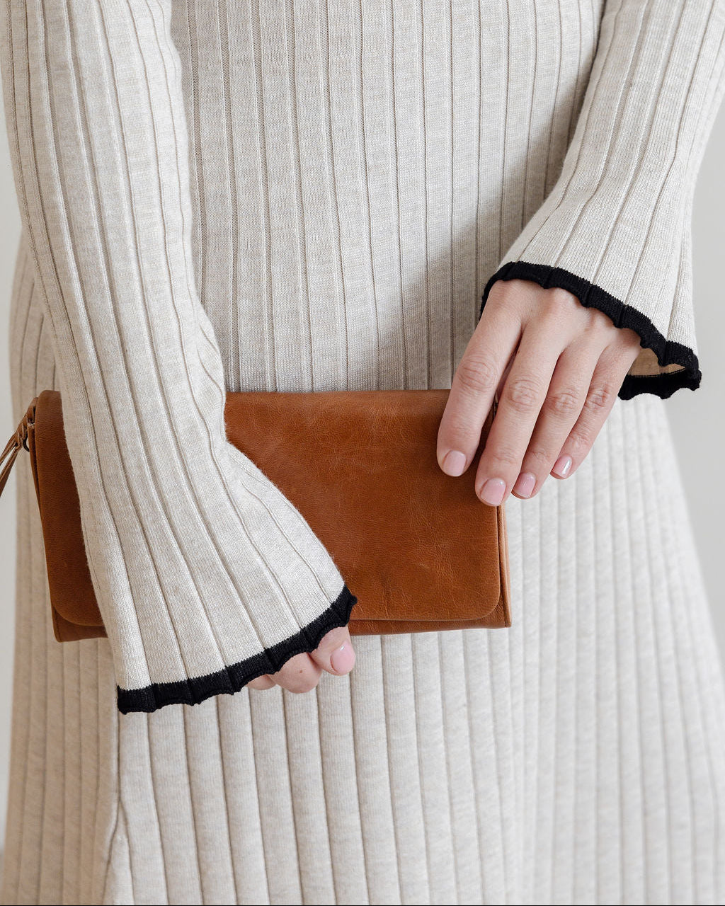 Person wearing a beige ribbed sweater holding a brown leather clutch.