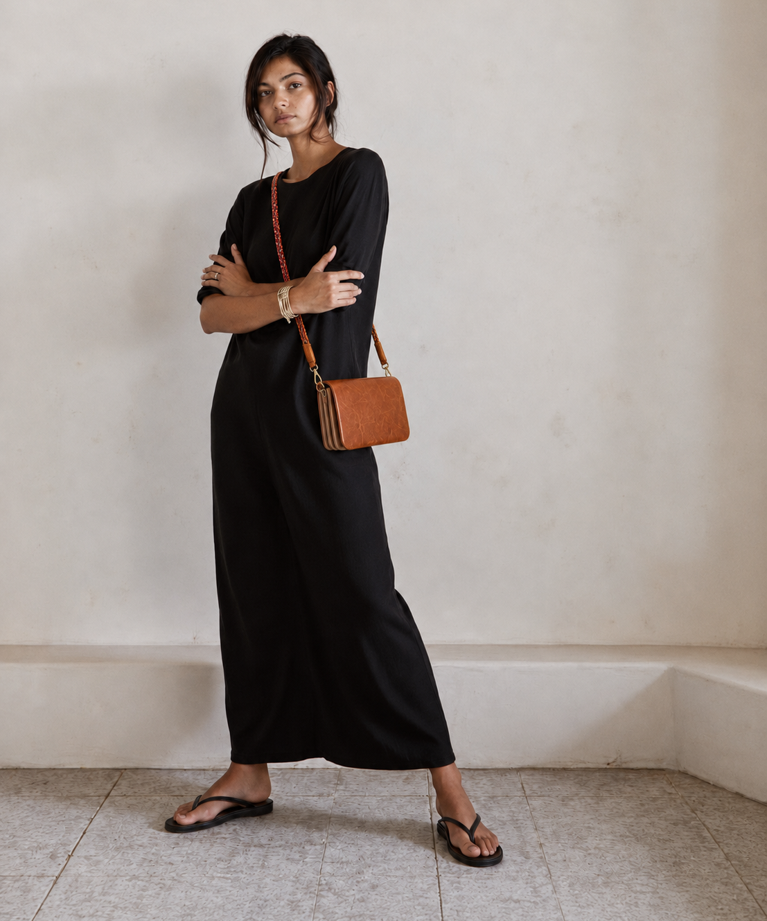 A woman stands in a minimalist room wearing a black long-sleeve maxi dress, black flip-flop sandals, and carrying a brown leather crossbody bag with a red chain strap.