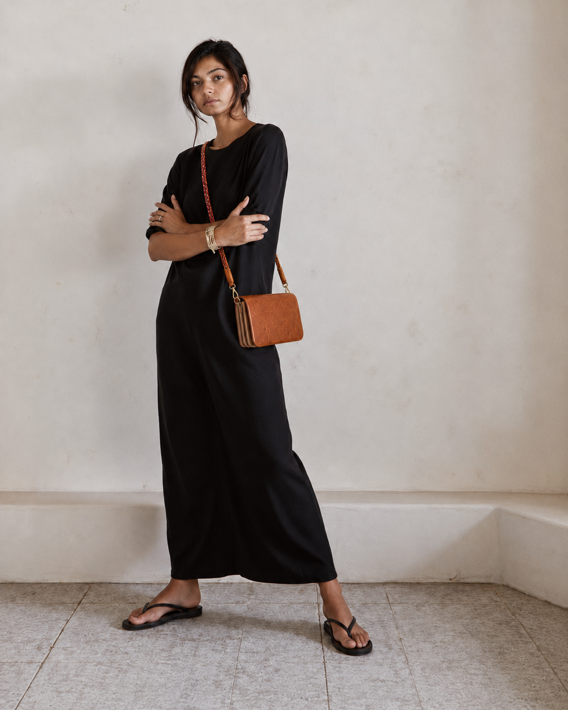 A woman stands in a minimalist room wearing a black long-sleeve maxi dress, black flip-flop sandals, and carrying a brown leather crossbody bag with a red chain strap.