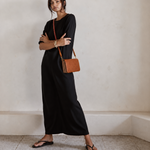 A woman stands in a minimalist room wearing a black long-sleeve maxi dress, black flip-flop sandals, and carrying a brown leather crossbody bag with a red chain strap.