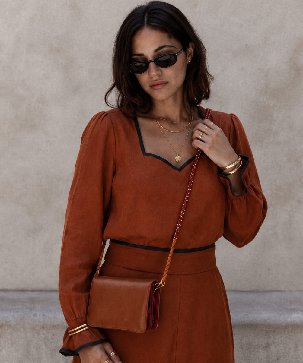 A woman wearing a rust-orange long-sleeve dress with black trim, black sunglasses, and gold jewelry, carrying a matching rust-brown leather crossbody bag against a textured beige wall.