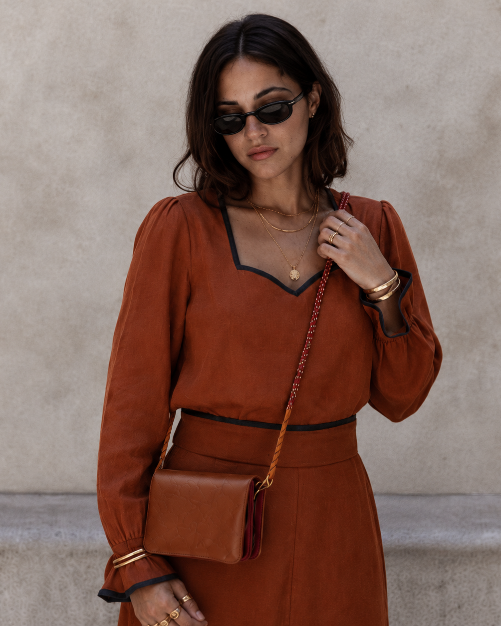 A woman wearing a rust-orange long-sleeve dress with black trim, black sunglasses, and gold jewelry, carrying a matching rust-brown leather crossbody bag against a textured beige wall.