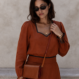 A woman wearing a rust-orange long-sleeve dress with black trim, black sunglasses, and gold jewelry, carrying a matching rust-brown leather crossbody bag against a textured beige wall.