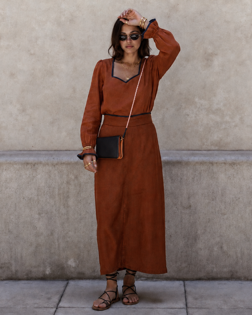 A woman stands against a textured beige wall wearing a rust-orange long-sleeve maxi dress with black trim, a black crossbody bag, and brown strappy sandals.