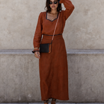 A woman stands against a textured beige wall wearing a rust-orange long-sleeve maxi dress with black trim, a black crossbody bag, and brown strappy sandals.