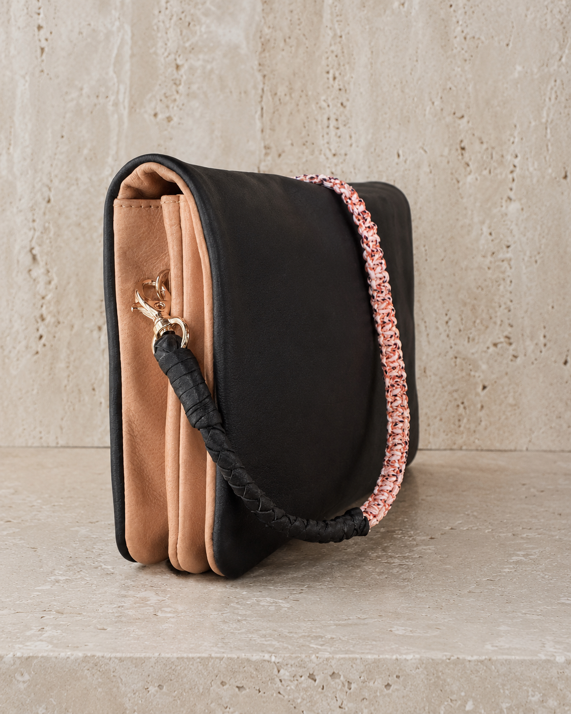 A structured black leather shoulder bag with a tan suede-like front panel, a woven black-and-pink strap, and a gold-tone clasp, displayed on a stone-textured surface.