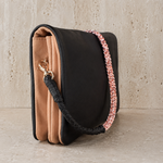 A structured black leather shoulder bag with a tan suede-like front panel, a woven black-and-pink strap, and a gold-tone clasp, displayed on a stone-textured surface.