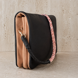 A structured black leather shoulder bag with a tan suede-like front panel, a woven black-and-pink strap, and a gold-tone clasp, displayed on a stone-textured surface.