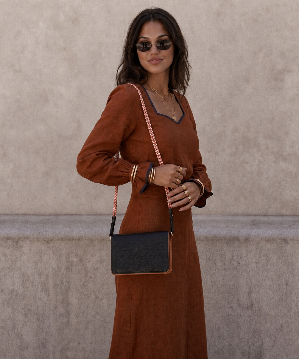 A woman wearing a rust-brown long-sleeve maxi dress with dark trim, styled with a black crossbody bag featuring a pink-and-white chain strap, standing against a textured beige wall.