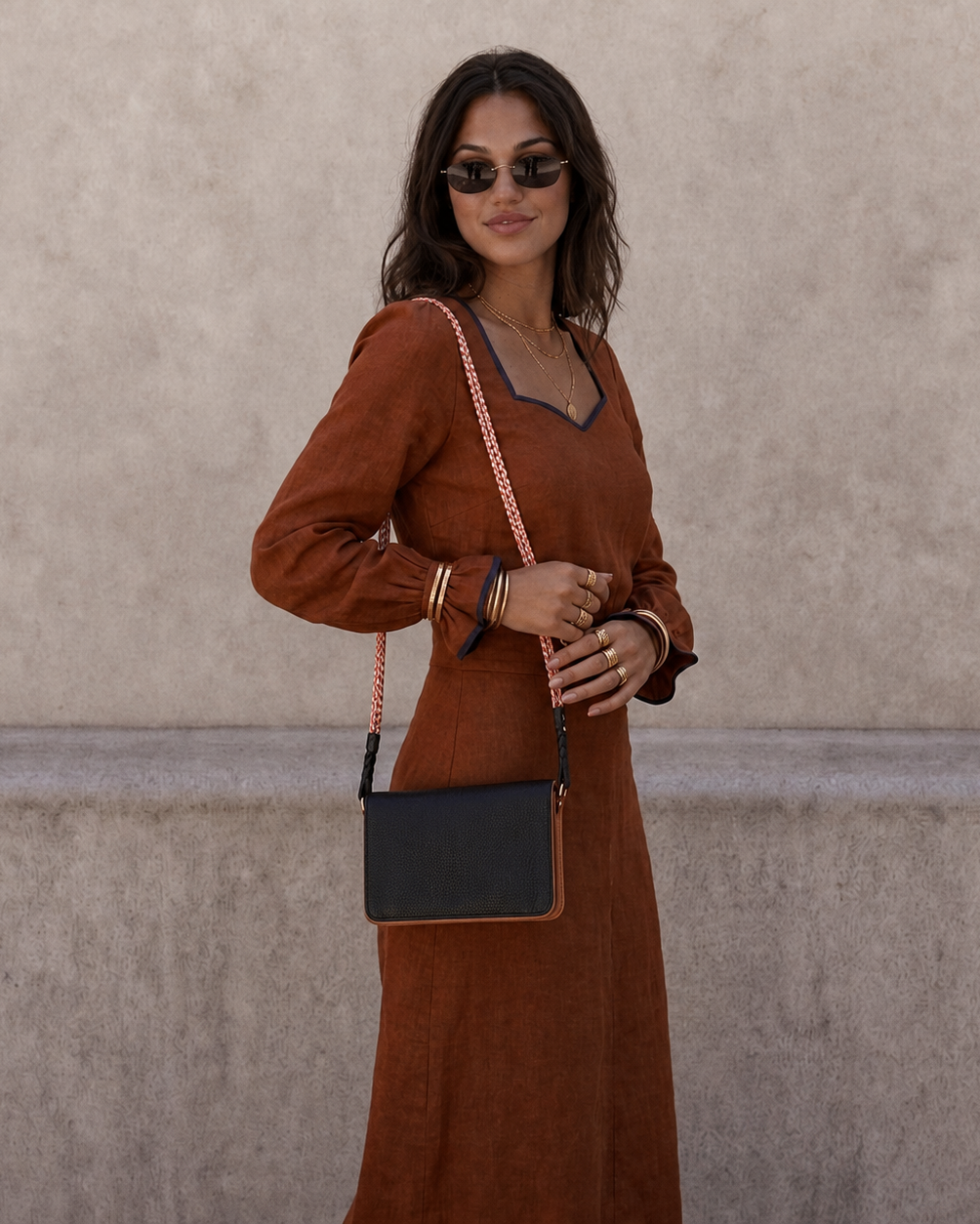 A woman wearing a rust-brown long-sleeve maxi dress with dark trim, styled with a black crossbody bag featuring a pink-and-white chain strap, standing against a textured beige wall.