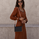 A woman wearing a rust-brown long-sleeve maxi dress with dark trim, styled with a black crossbody bag featuring a pink-and-white chain strap, standing against a textured beige wall.