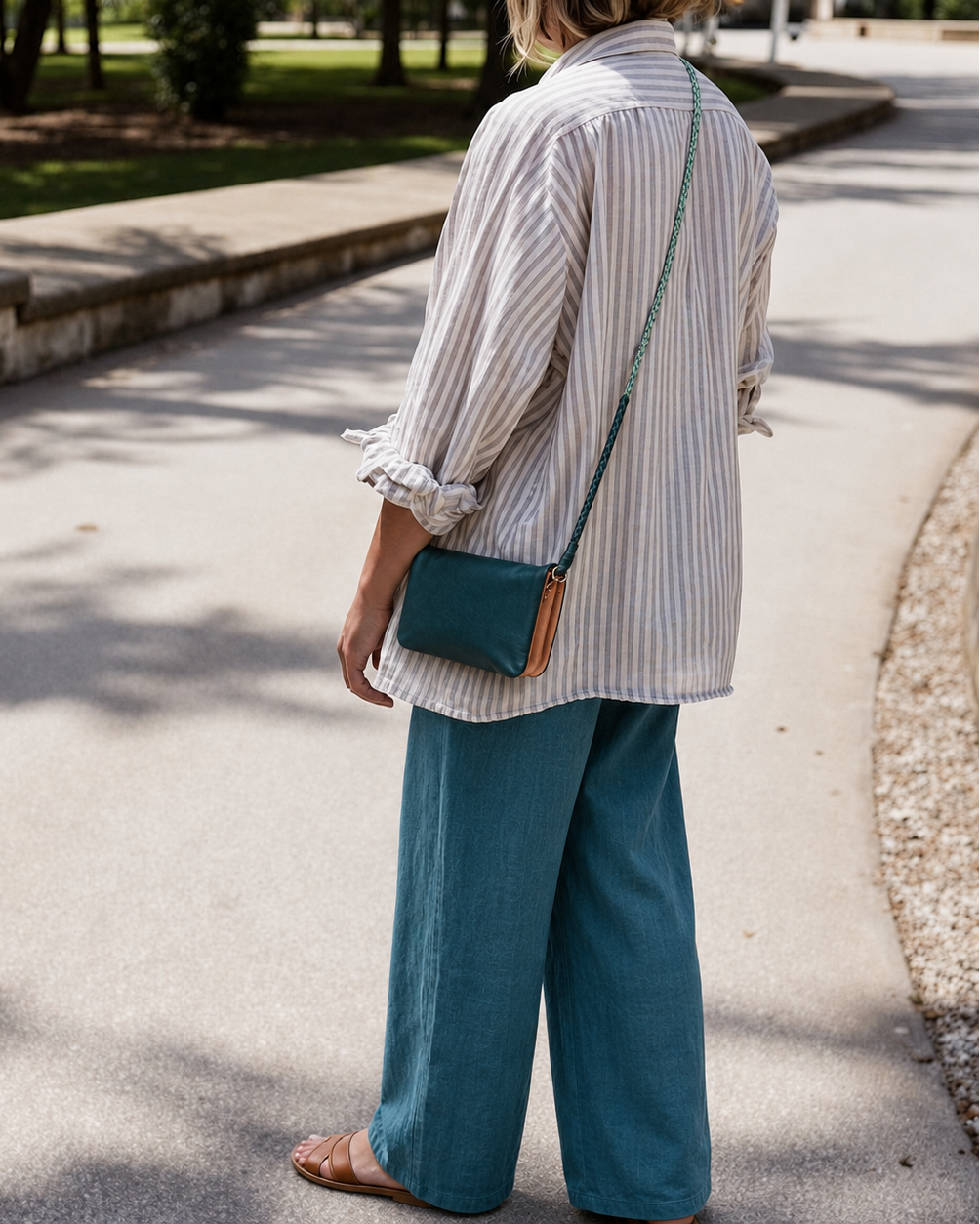 A woman standing on a paved outdoor path, shown from the back, wearing a white and blue vertically striped button-up shirt, teal wide-leg pants, and carrying a small teal crossbody bag with a tan leather accent.