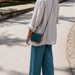 A woman standing on a paved outdoor path, shown from the back, wearing a white and blue vertically striped button-up shirt, teal wide-leg pants, and carrying a small teal crossbody bag with a tan leather accent.