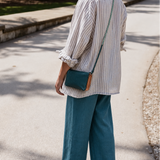 A woman standing on a paved outdoor path, shown from the back, wearing a white and blue vertically striped button-up shirt, teal wide-leg pants, and carrying a small teal crossbody bag with a tan leather accent.