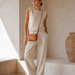 A woman stands in a minimalist beige interior wearing a sleeveless cream knit top and matching wide-leg trousers, with a small tan crossbody bag and flat sandals.