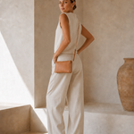 A woman stands in a minimalist beige interior wearing a sleeveless cream top and matching wide-leg pants, with a small tan crossbody bag and flat sandals.