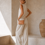 A woman stands in a minimalist beige interior wearing a sleeveless cream top and matching wide-leg pants, with a small tan crossbody bag and flat sandals.