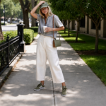 A woman stands on a tree-lined sidewalk wearing a light sage green short-sleeve button-up shirt, off-white cropped wide-leg pants, olive green sneakers, and carrying a small olive green shoulder bag.