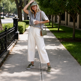 A woman stands on a tree-lined sidewalk wearing a light sage green short-sleeve button-up shirt, off-white cropped wide-leg pants, olive green sneakers, and carrying a small olive green shoulder bag.