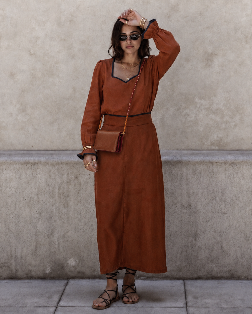 A woman stands against a textured concrete wall wearing a rust-brown long-sleeve maxi dress with a V-neckline, black trim, and a matching belt. She is accessorized with a small brown crossbody bag, sunglasses, bracelets, and brown strappy sandals.