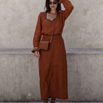 A woman stands against a textured concrete wall wearing a rust-brown long-sleeve maxi dress with a V-neckline, black trim, and a matching belt. She is accessorized with a small brown crossbody bag, sunglasses, bracelets, and brown strappy sandals.