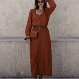 A woman stands against a textured concrete wall wearing a rust-brown long-sleeve maxi dress with a V-neckline, black trim, and a matching belt. She is accessorized with a small brown crossbody bag, sunglasses, bracelets, and brown strappy sandals.
