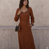 A woman wearing a rust-brown long-sleeve midi dress with a square neckline and black trim, standing against a textured beige wall. She is carrying a small black and brown shoulder bag.
