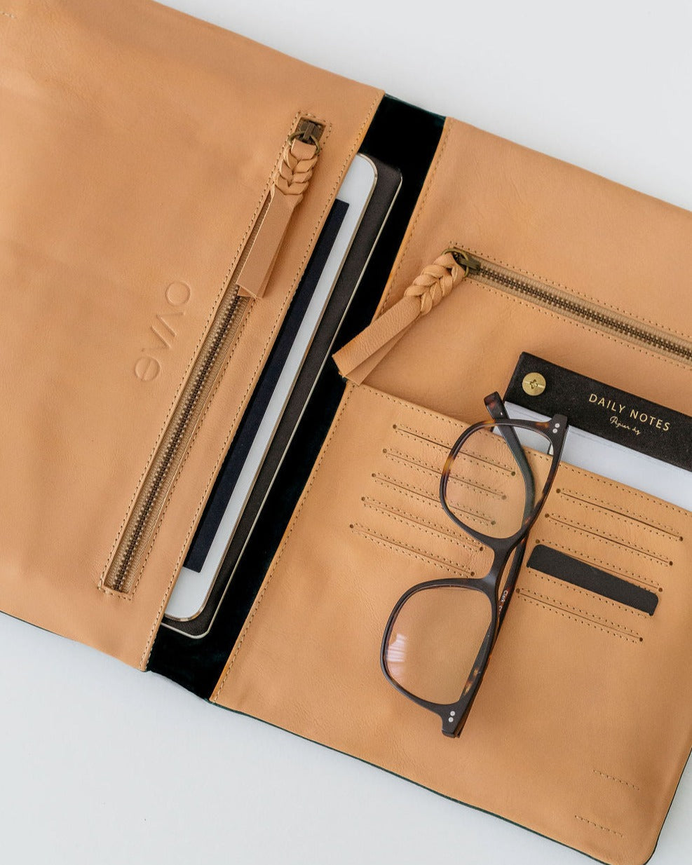 A black leather organiser with multiple compartments, displaying slots for cards, a pocket for a tablet or phone, and zip compartments. It is laid open to show the interior, with an eyewear case placed on one of the pockets.