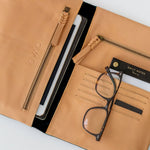 A black leather organiser with multiple compartments, displaying slots for cards, a pocket for a tablet or phone, and zip compartments. It is laid open to show the interior, with an eyewear case placed on one of the pockets.